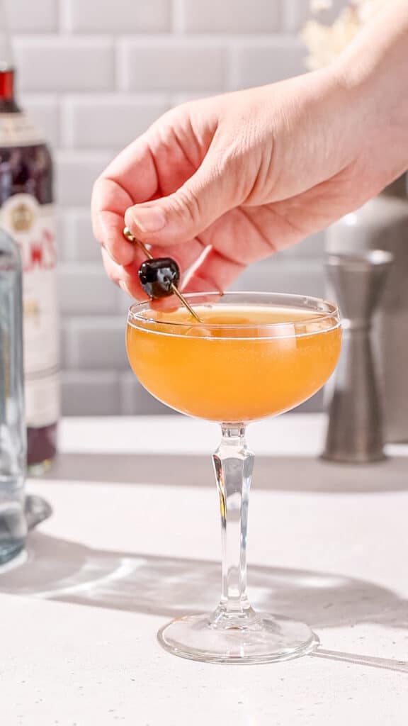 Hand adding a maraschino cherry on a pick as a garnish to a cocktail in a coupe glass.