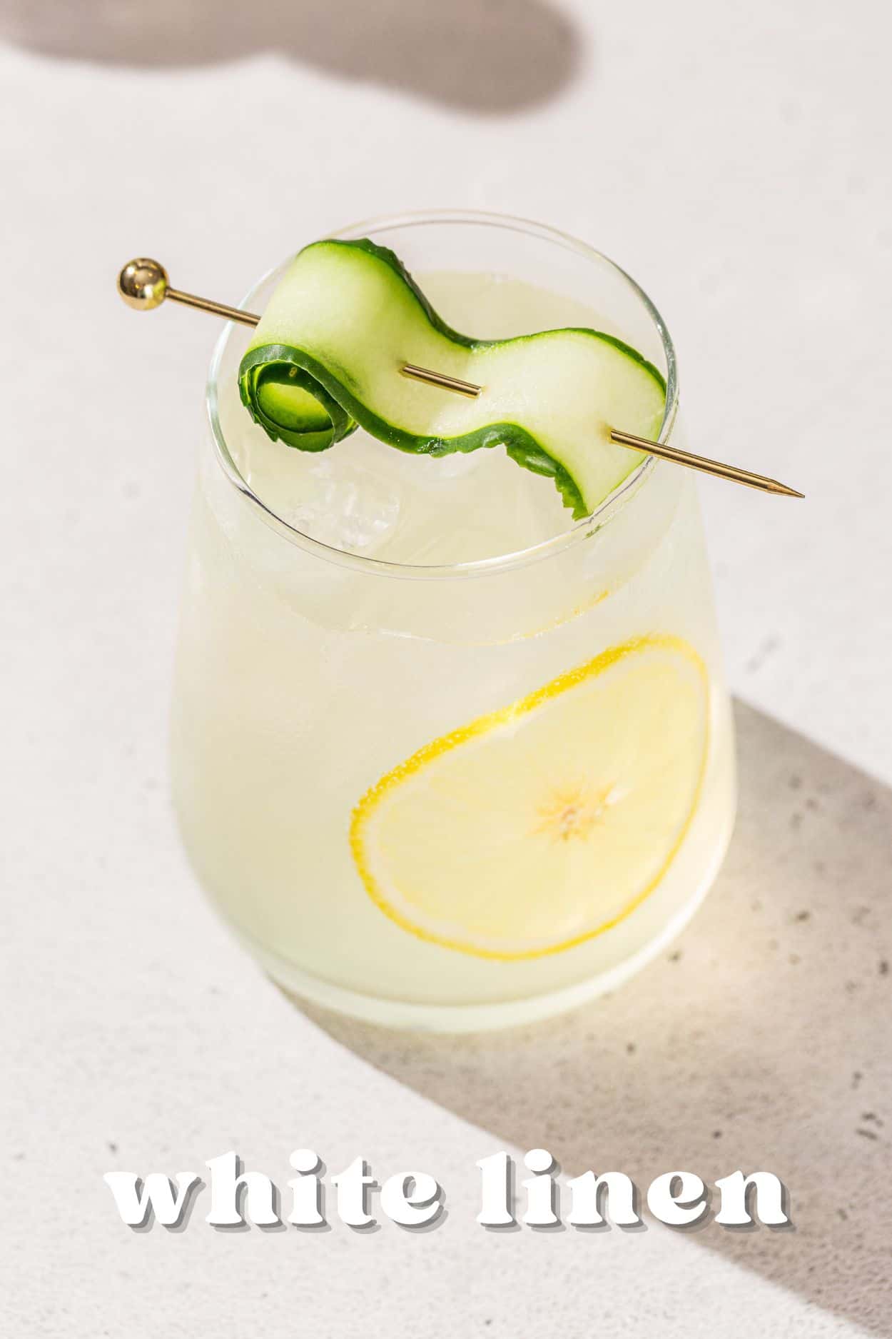 Overhead view of a refreshing-looking White Linen cocktail with a cucumber and lemon garnish. Text at the bottom says “Whte Linen”.