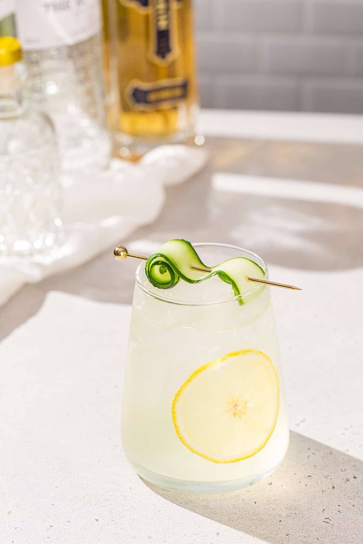 White Linen cocktail with a cucumber and lemon garnish in a stemless wine glass on a countertop. A bottle of gin and a bottle of St Germain elderflower liqueur are in the background.