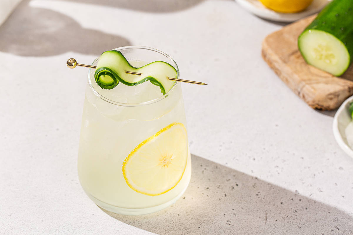 White Linen cocktail with a cucumber ribbon and lemon slice garnish in a stemless wine glass, sitting on a countertop. A sliced cucumber is to the right on a cutting board.