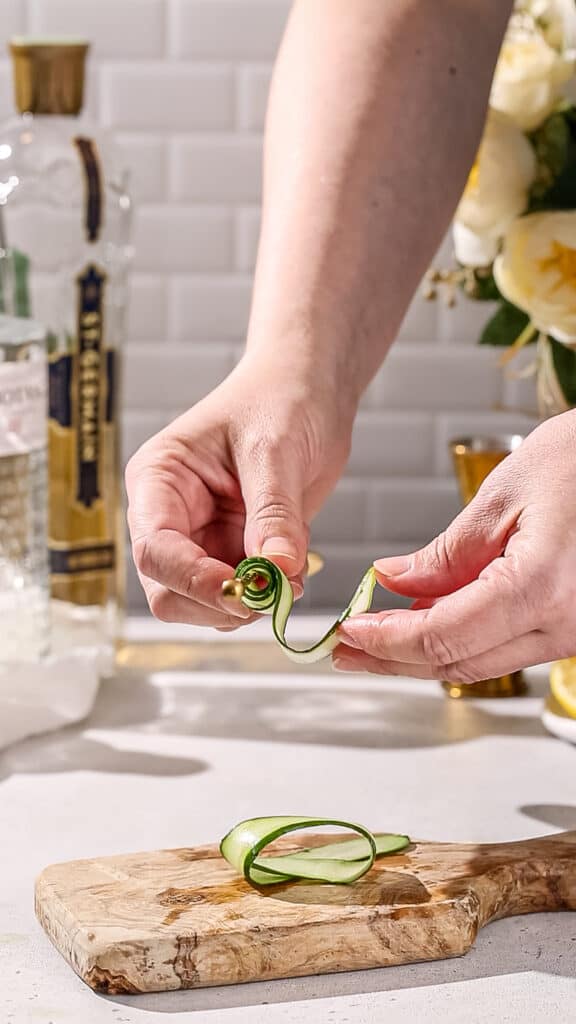 Hands curling up a cucumber ribbon for a drink garnish.