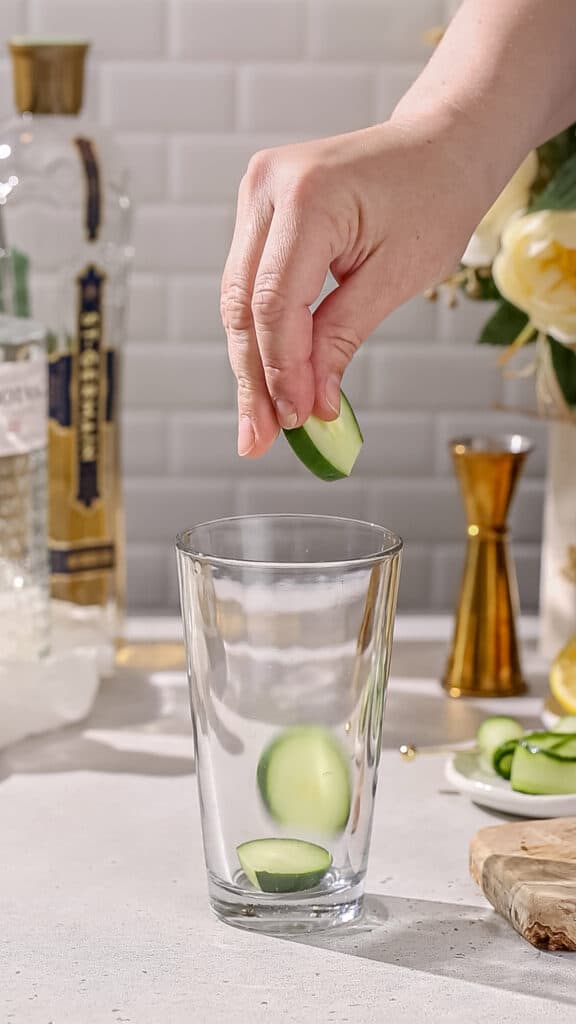 Hands dropping cucumber slices into a cocktail shaker.