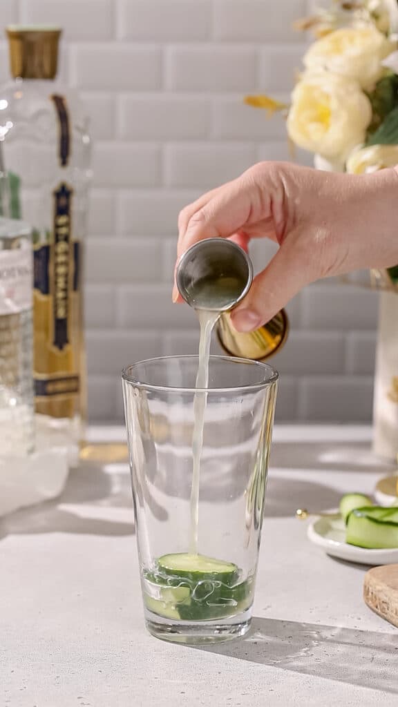 Hand adding lemon juice to a cocktail shaker that has cucumber in it.