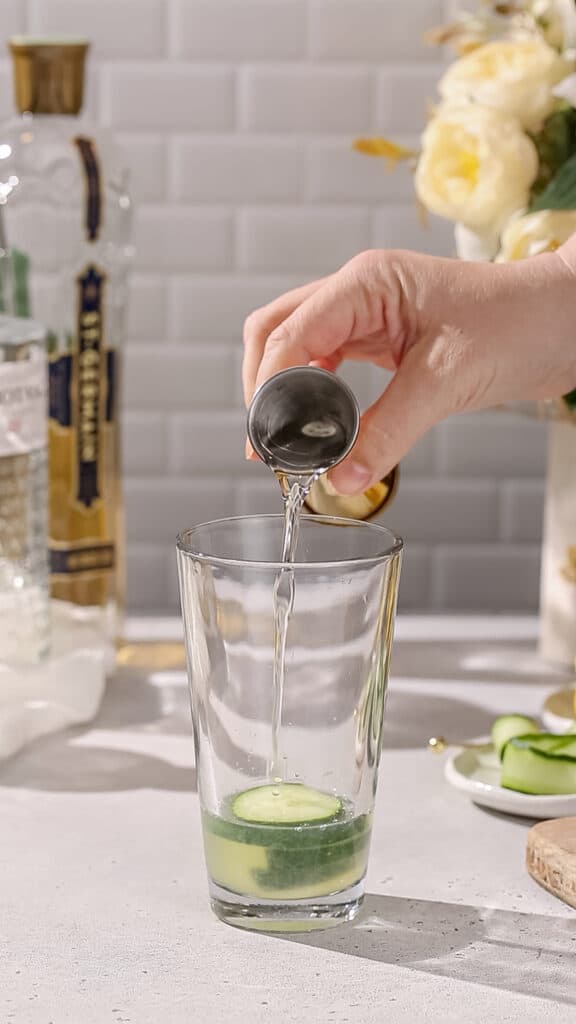 Hand adding simple syrup to a cocktail shaker with cucumbers.