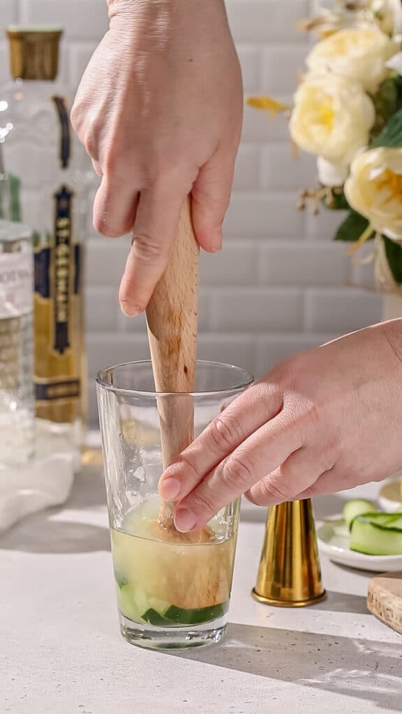 Hand using a wooden muddler to muddle cucumbers in a cocktail shaker.
