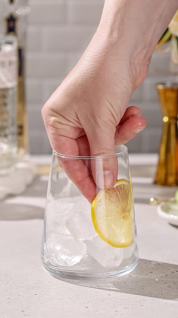 Hand adding a lemon slice to the side of a cocktail serving glass.