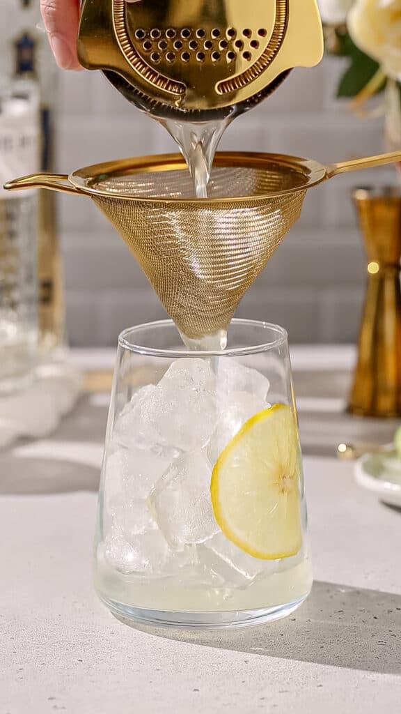 Cocktail being strained into a serving glass that has a lemon slice on the side and is filled with ice.