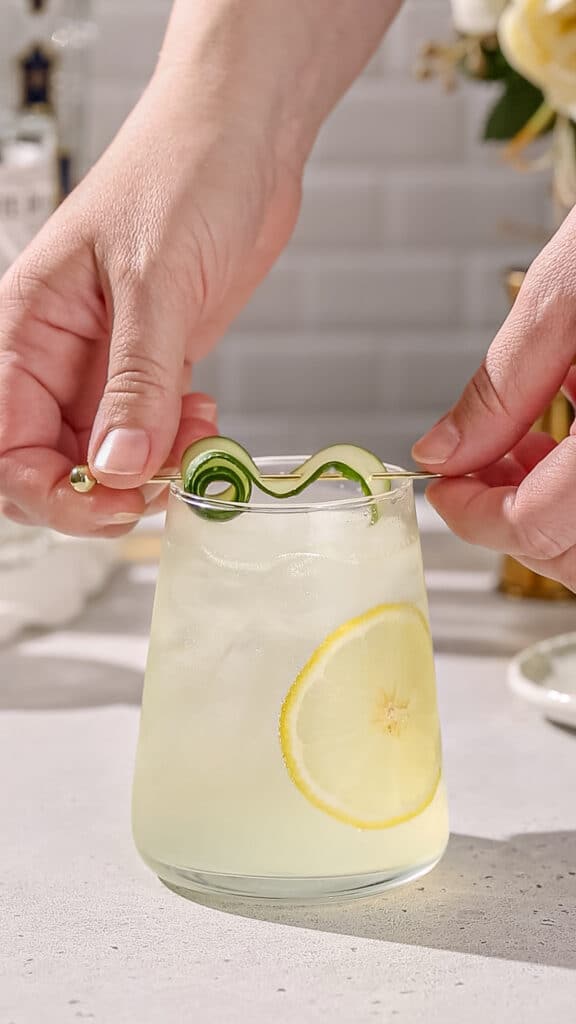 Hands adding a cucumber ribbon on a cocktail pick to the top of a cocktail glass that has a lemon slice on the side.