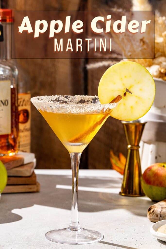 Side view of an Apple Cider Martini on a countertop. The drink has a cinnamon sugar rim, a cinnamon stick and an apple slice garnish. In the background are some ingredients and bar tools. Text at the top says “Apple Cider Martini” in bold letters.