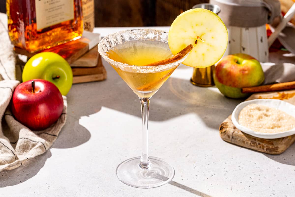 Slightly overhead view of an Apple Cider Martini on a countertop. The drink is in a Martini glass and has a cinnamon stick and apple slice garnish. In the background are some apples, cinnamon sugar, Amaretto and vodka.