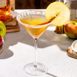 Slightly overhead view of an Apple Cider Martini on a countertop. The drink is in a Martini glass and has a cinnamon stick and apple slice garnish. In the background are some apples, cinnamon sugar, Amaretto and vodka.