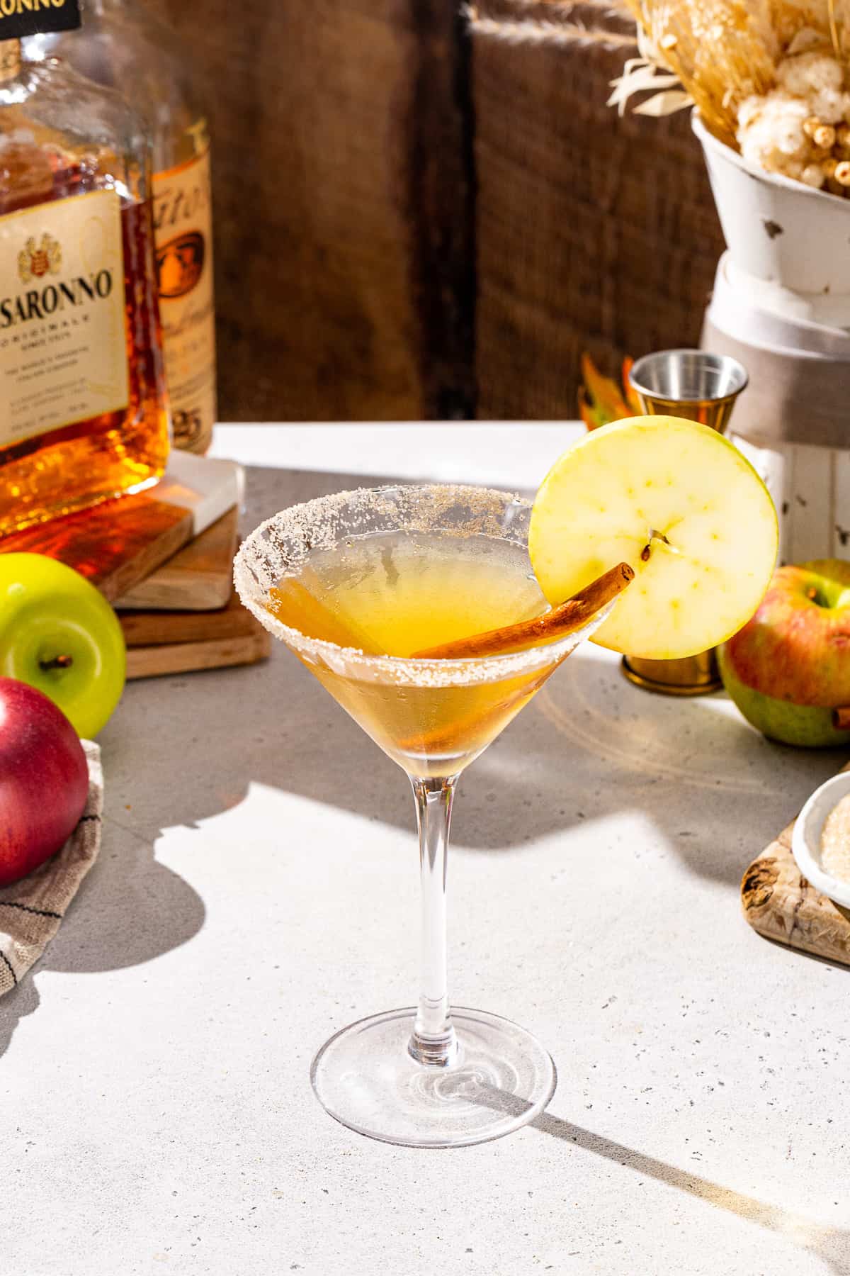 Apple Cider Martini on a countertop with a cinnamon stick and apple slice garnish. The drink has a cinnamon sugar rim. In the background are some apples, vodka and cinnamon sugar.