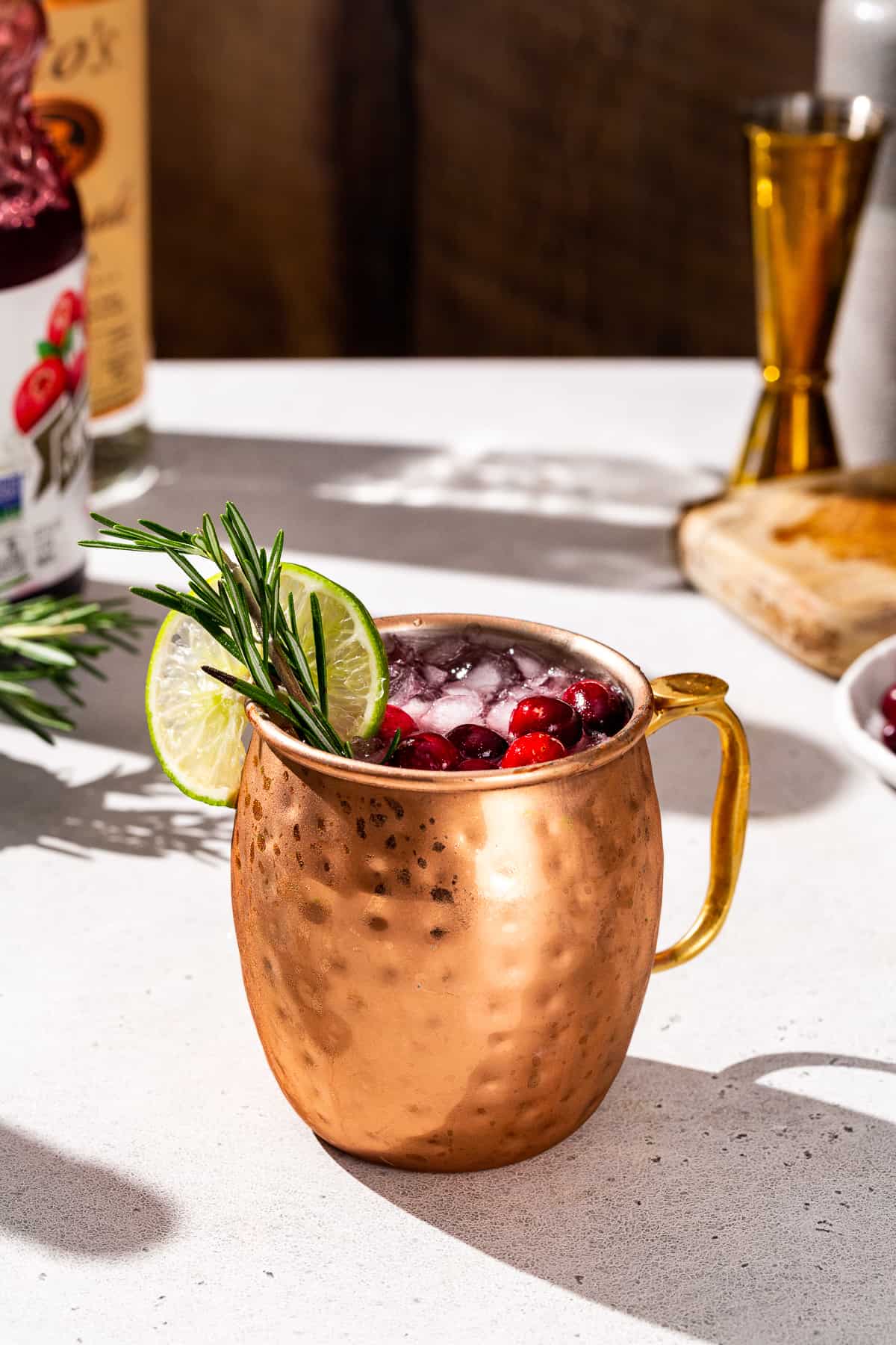 Side view of a Christmas Mule cocktail in a copper mug. The drink has cranberries floating in it along with a rosemary and lime garnish. Ingredients and bar tools are seen in the background.