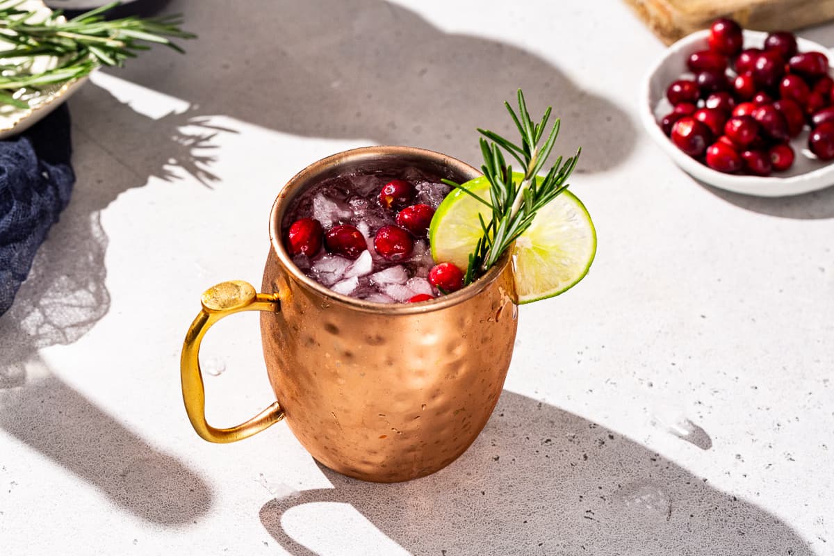 Christmas Mule cocktail in a copper mug on a countertop. The drink is garnished with fresh cranberries, rosemary and a lime slice.