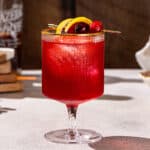 Side view of a bright red Cranberry whiskey Sour cocktail with a cranberry and lemon peel garnish. The drink is in a modern style cocktail glass with a short stem and a gold rim.