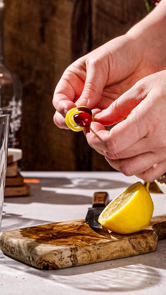 Hands wrapping a lemon peel around cranberries on a cocktail pick.