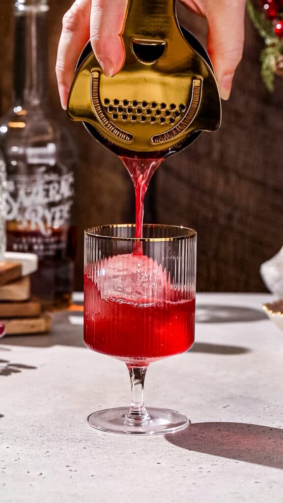 Hand using a cocktail shaker and Hawthorne strainer to pour red liquid into a cocktail glass that has a large ice sphere in it.