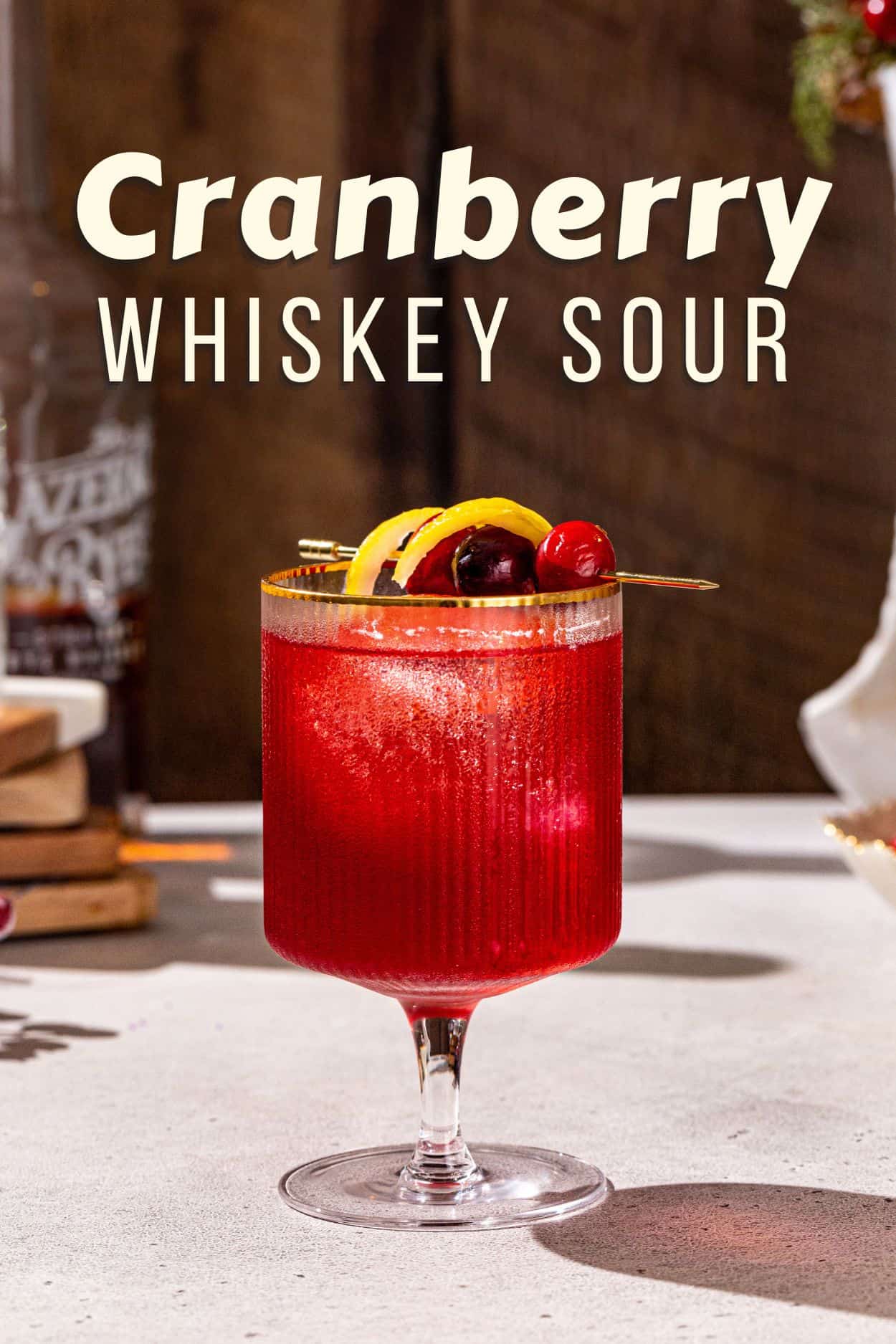 Side view of a Cranberry Whiskey Sour on a countertop. In the background a bottle of rye whiskey is visible. The drink itself is bright red with a cranberry and lemon peel garnish. Bold text at the top says “Cranberry whiskey sour”.