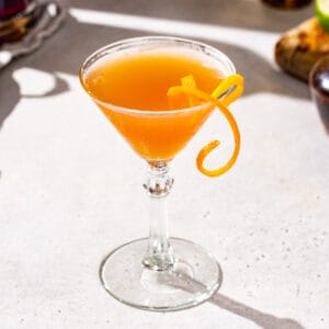 Lion’s Tail cocktail in a martini glass on a white countertop. The drink is orange in color and has a long curly strip of orange peel as the garnish.