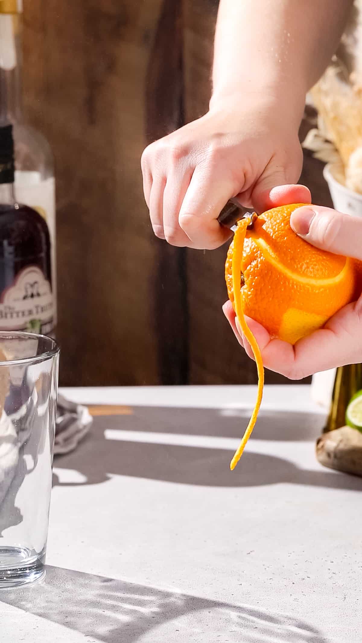 Hands using a channel knife to cut a long strip of orange peel.