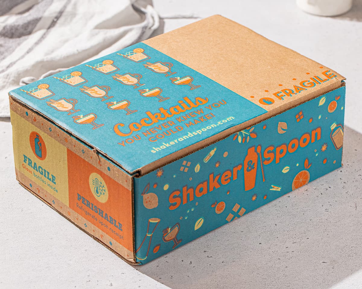 Shaker & Spoon cardboard box on a countertop. The box is brown with blue and orange printing on it. It has the Shaker & Spoon logo on the side, and the top says “Cocktails you never knew you could make”. It has Perishable and Fragile printed on it as well.