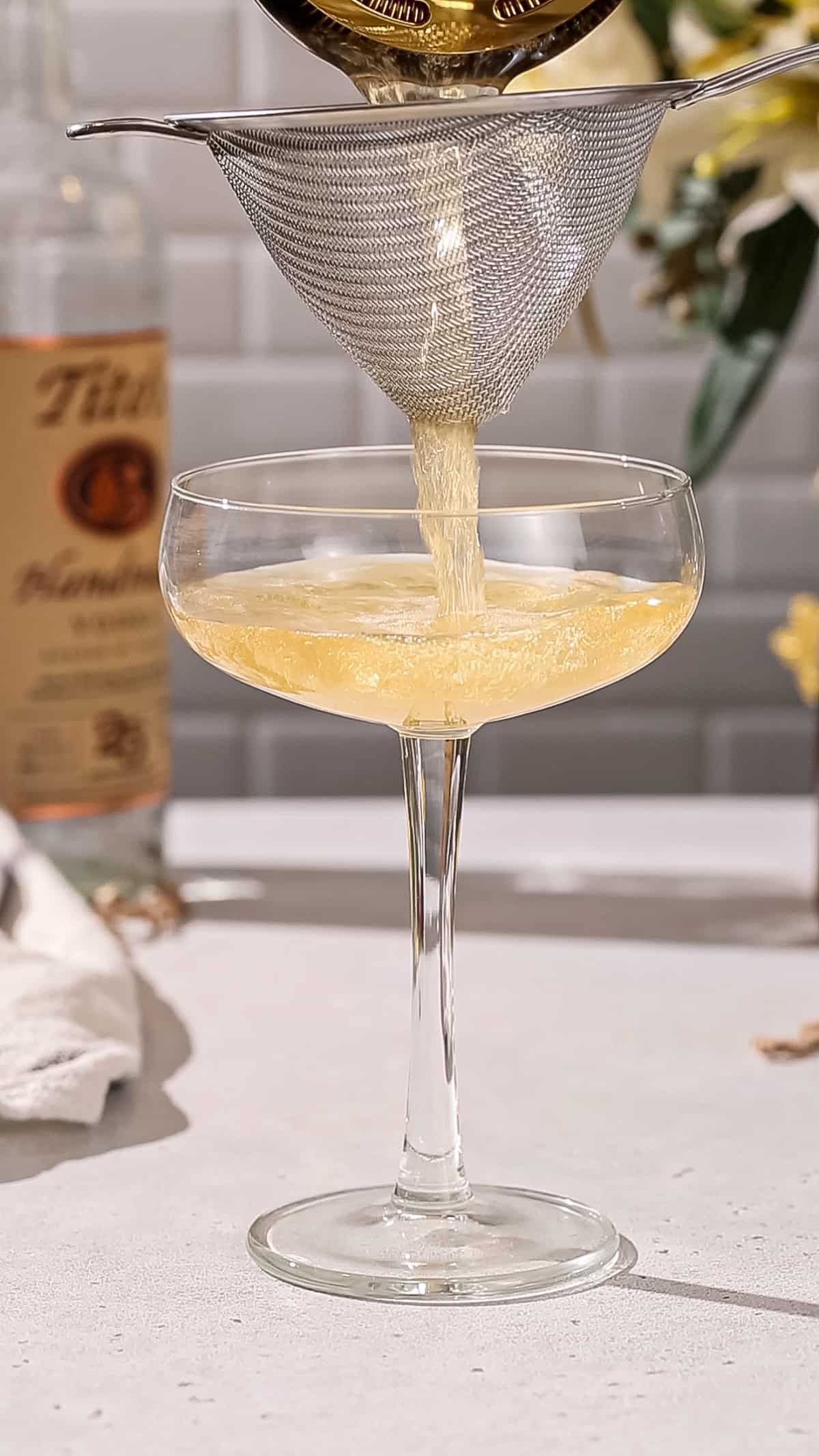 Cocktail being poured into a coupe glass through a regular strainer and a fine mesh strainer.