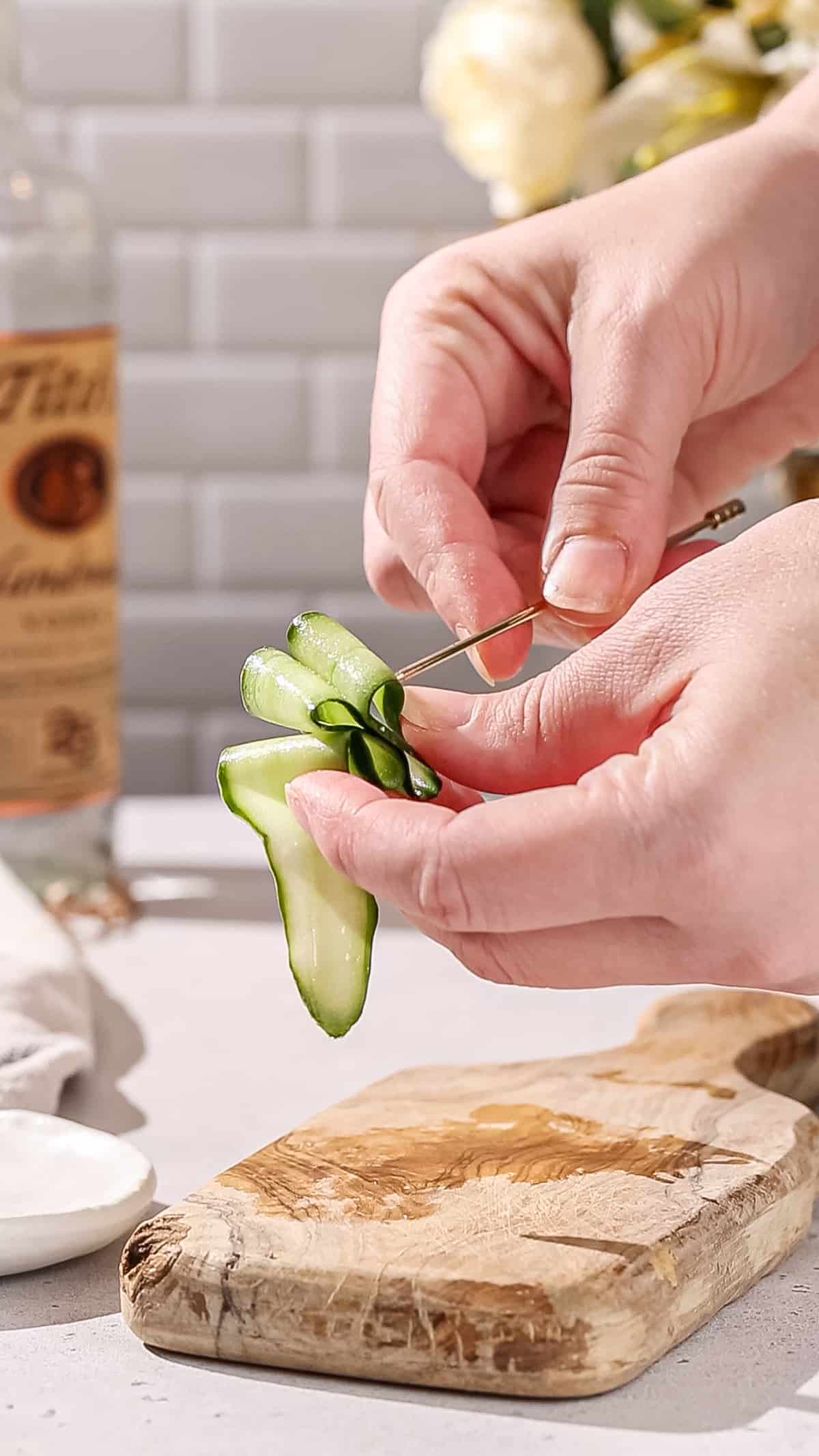 Hands adding a long strip of cucumber to a cocktail pick in a zigzag ribbon style.