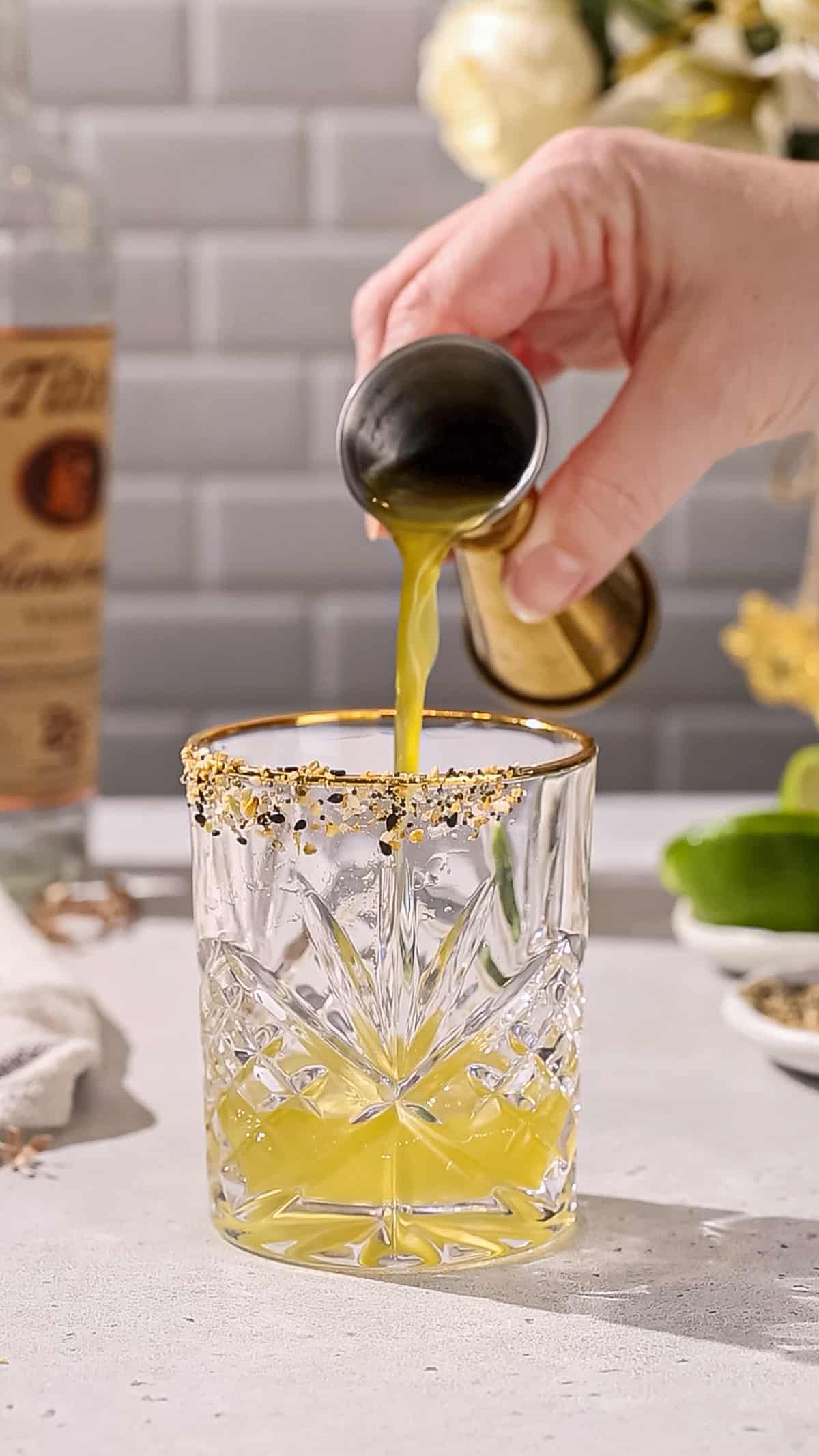 Hand adding a green shrub liquid to a cocktail glass.