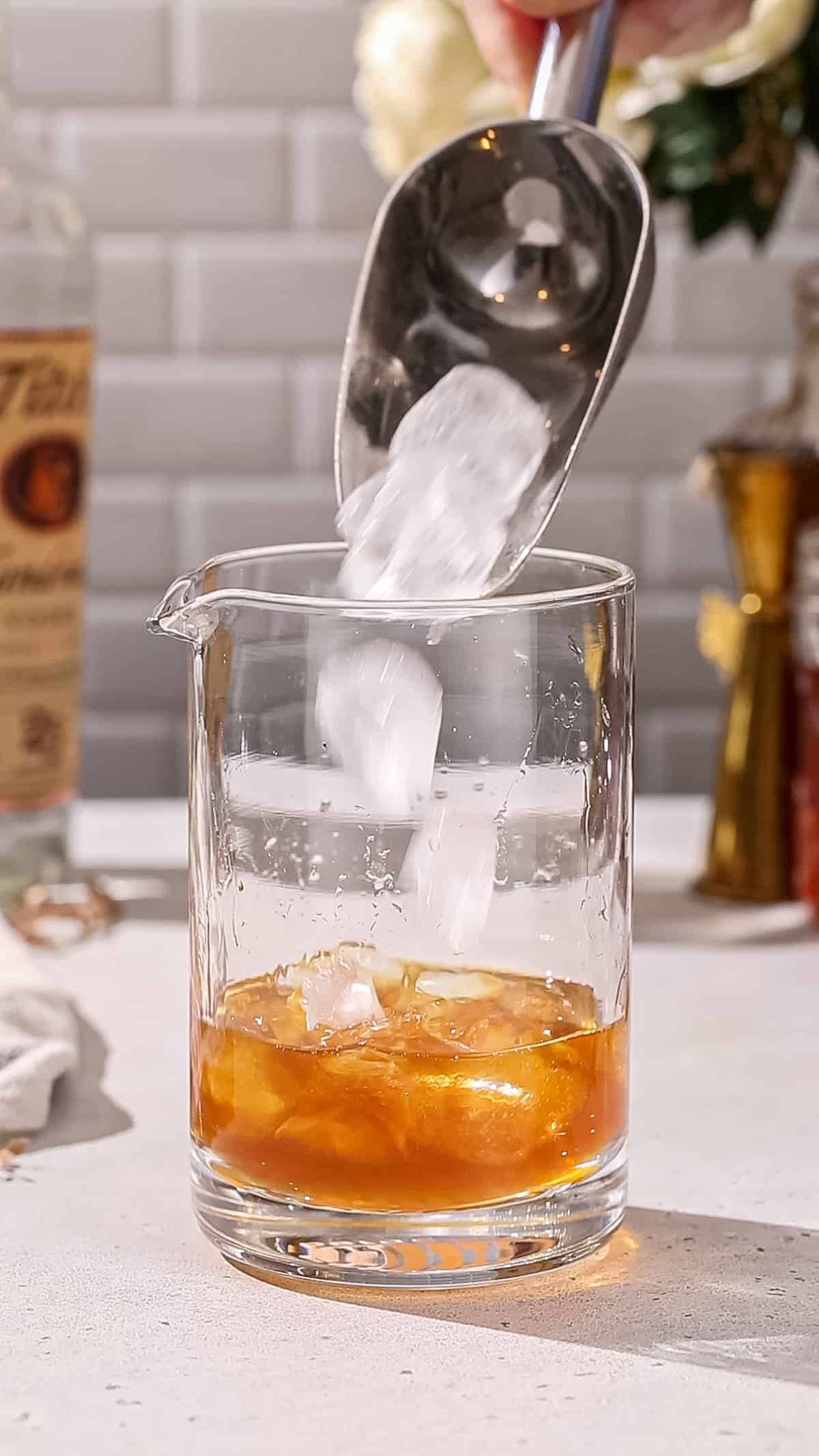 Hand using an ice scoop to add ice to a cocktail mixing glass with amber liquid in it.