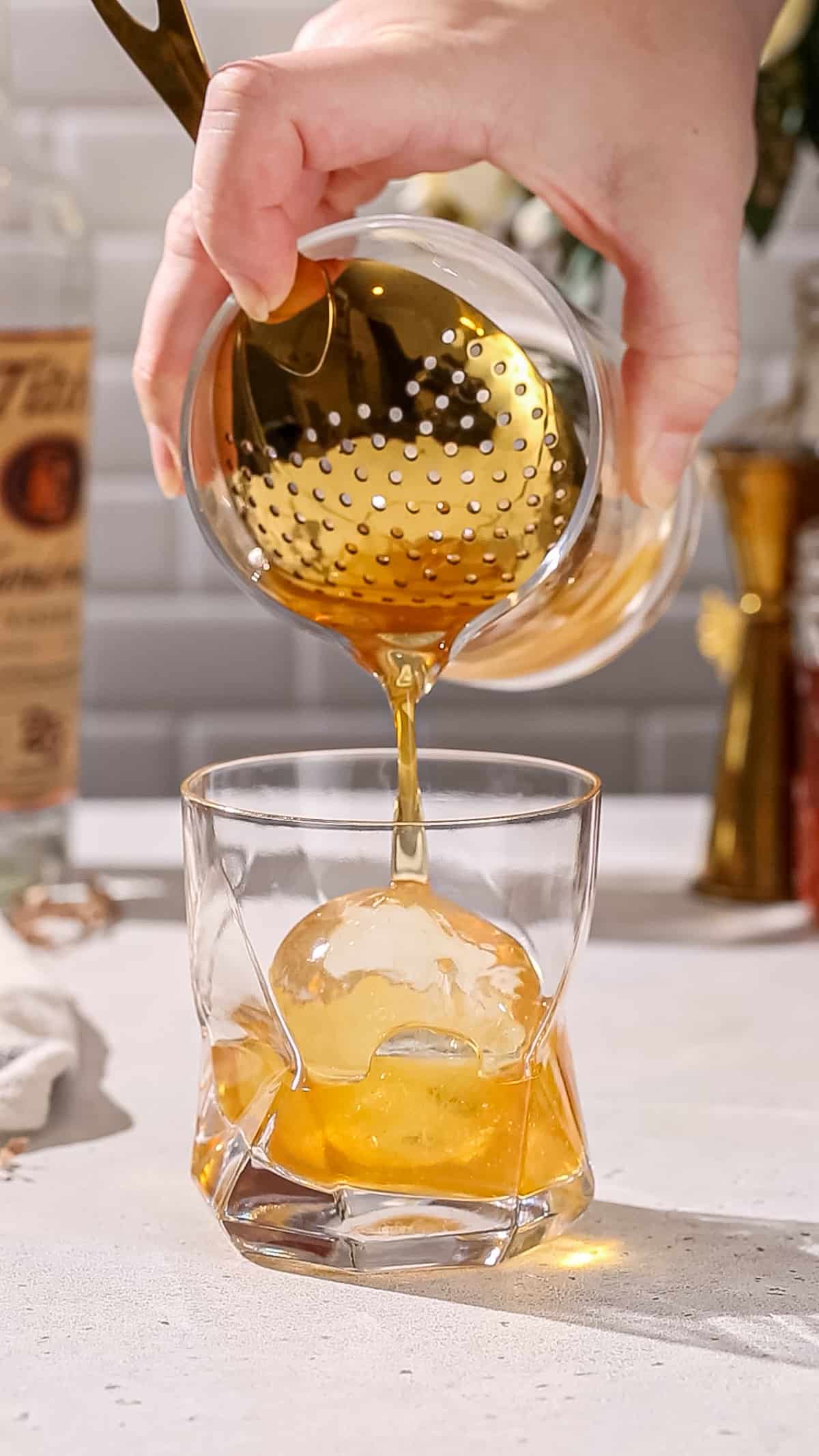 Hand using a mixing glass and a Julep strainer to strain an amber colored cocktail over top of the ice sphere.