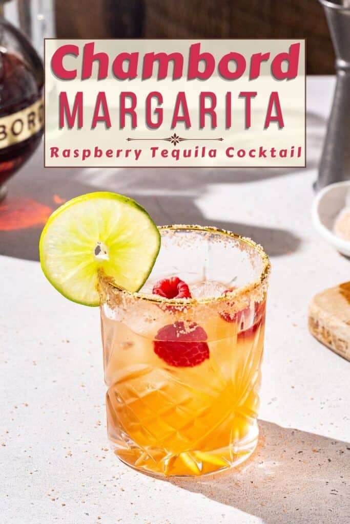 Slighty overhead view of a Chambord raspberry margarita. The drink is in a gold-rimmed glass that also has salt on the rim. It has a cut lime and raspberries as garnish. A bottle of Chambord liqueur can be seen off to the left. Text overlay above the drink says “Chambord Margarita - Raspberry Tequila cockail”.