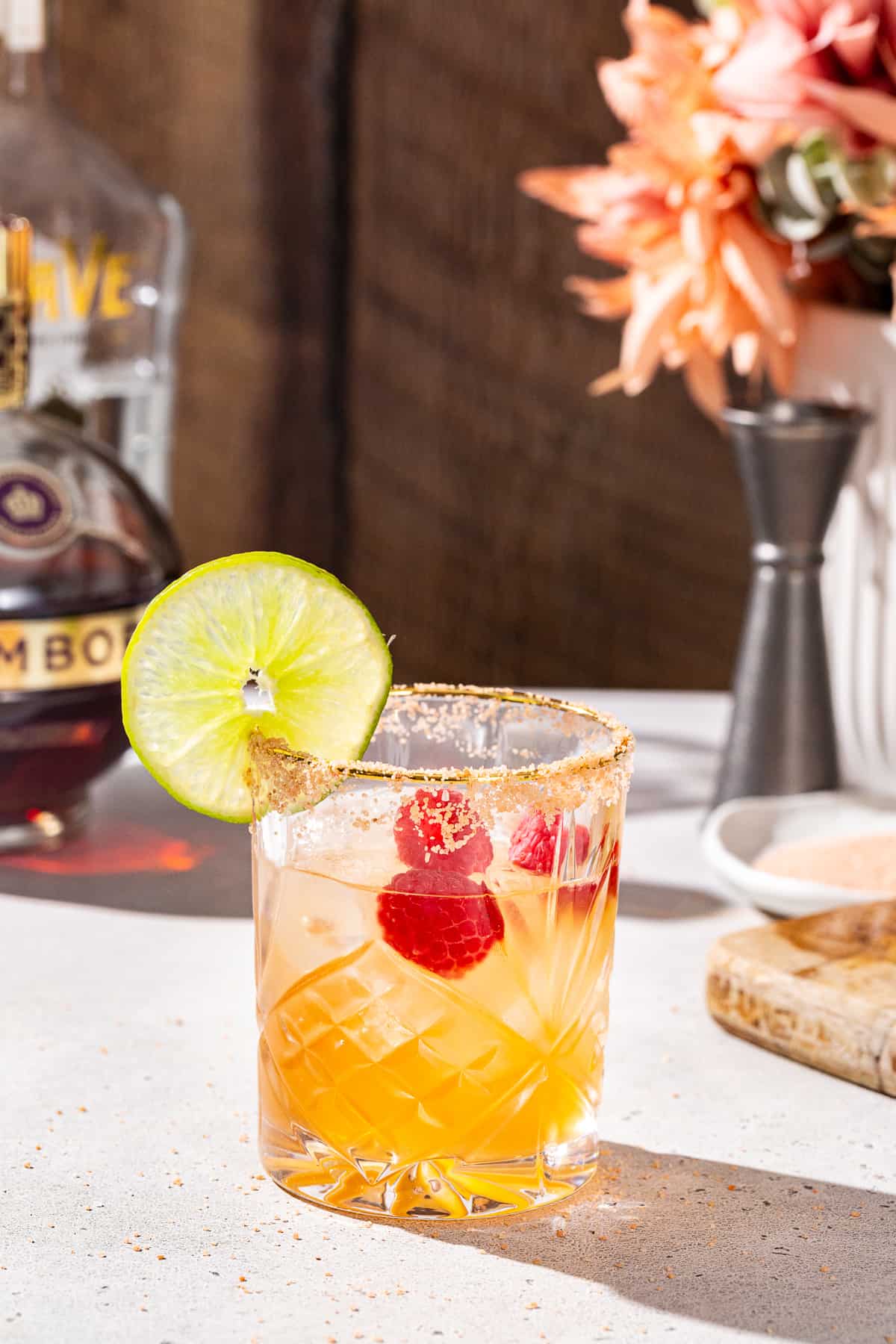 Chambord Margarita on a countertop with a bottle of Chambord, tequila, a jigger and some salt in the background. The drink is in an old fashioned glass and has fresh raspberries and a slice of lime as a garnish.