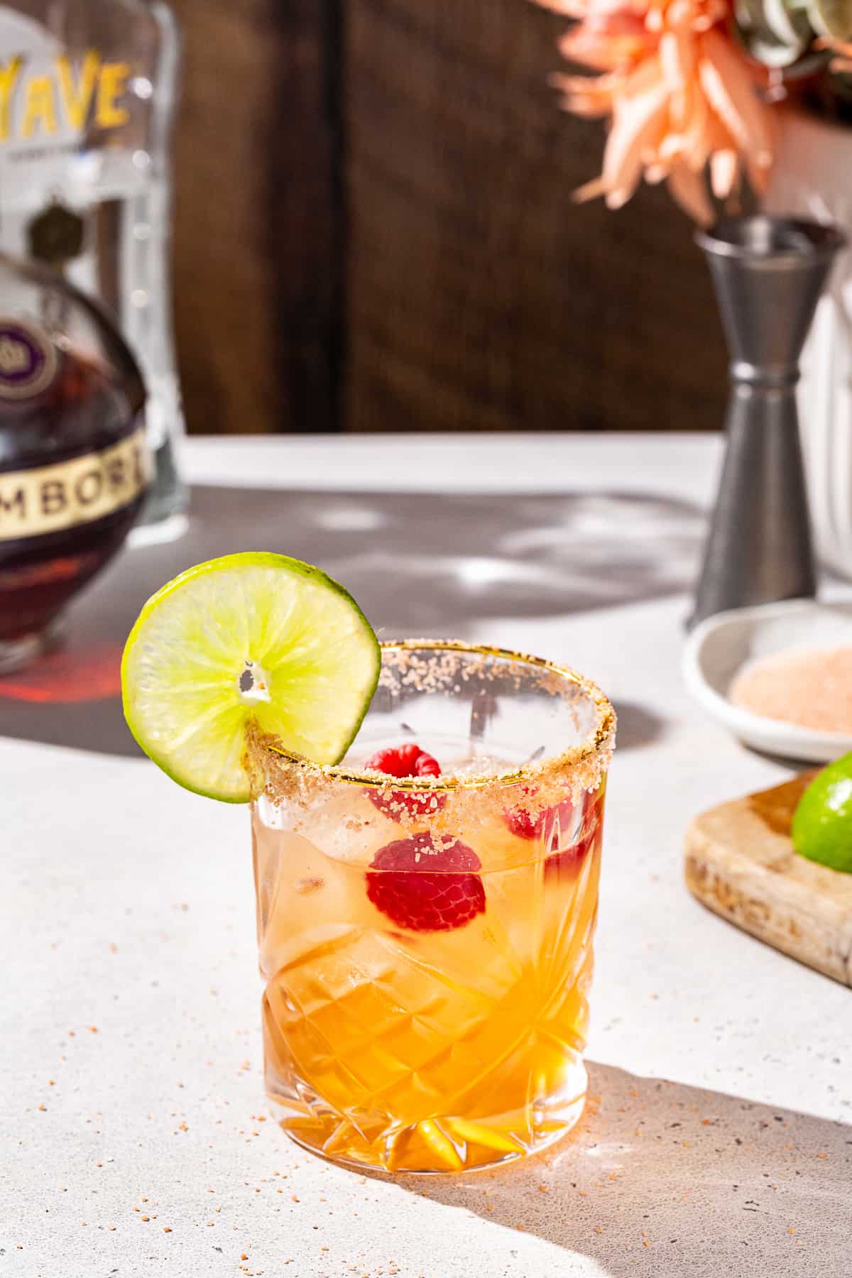 Slightly overhead view of a Chambord Margarita on a countertop with a bottle of Chambord, tequila blanco, a jigger and a cut lime in the background. The drink is in an old fashioned glass and has fresh raspberries and a slice of lime as a garnish.