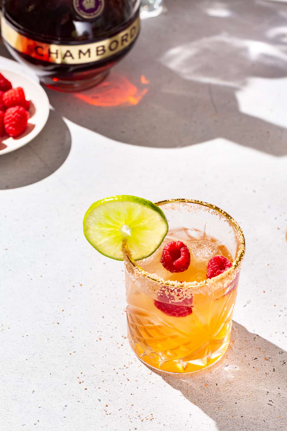 Overhead view of a Chambord raspberry margarita. The drink is in a gold-rimmed glass that also has salt on the rim. It has a cut lime and raspberries as garnish. A dish of fresh raspberries is seen in the background along with a bottle of Chambord liqueur.