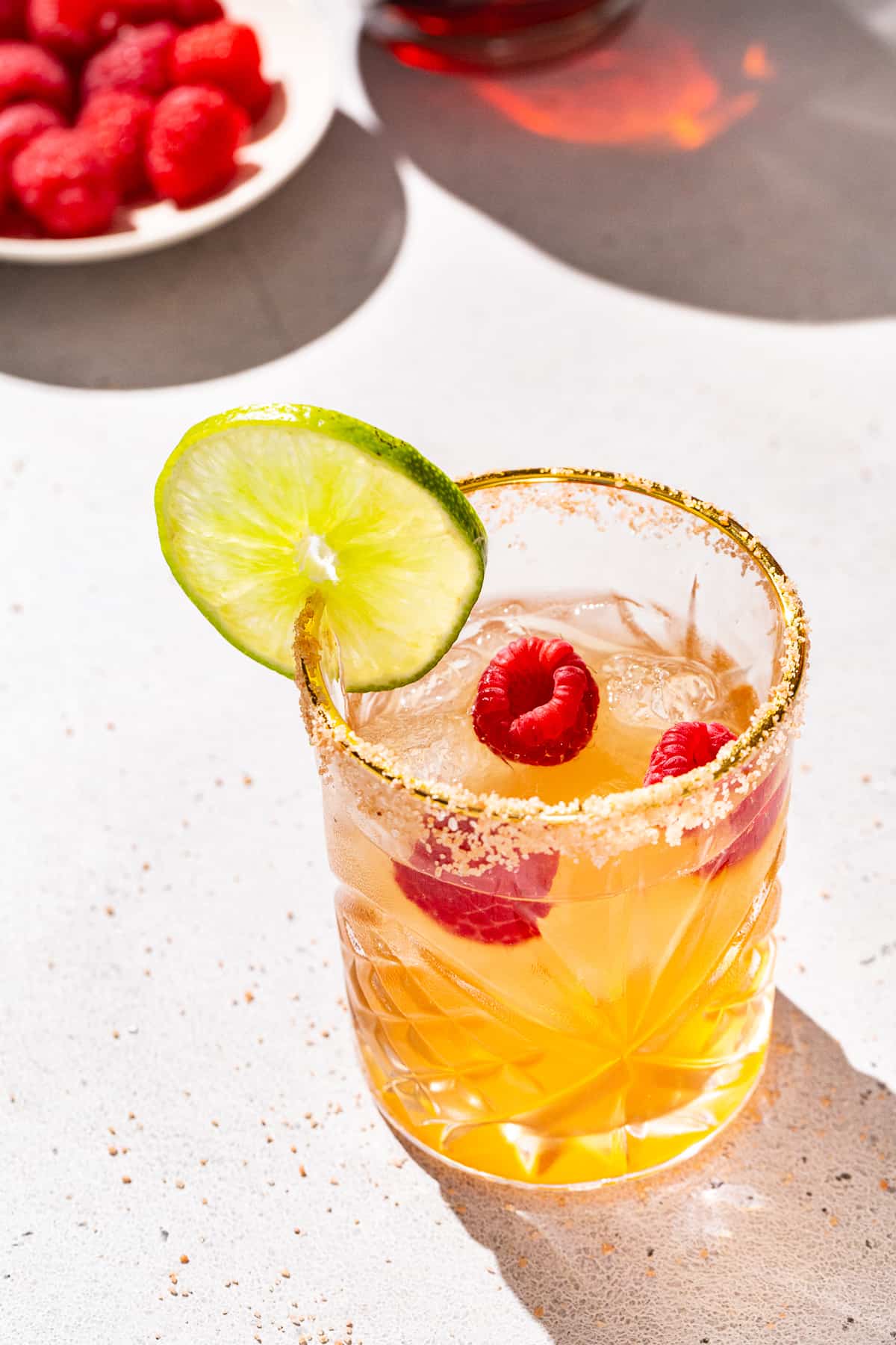 Overhead view of a Chambord raspberry margarita. The drink is in a gold-rimmed glass that also has salt on the rim. It has a cut lime and raspberries as garnish. A dish of fresh raspberries is seen in the background along with a bottle of Chambord liqueur.