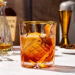 Close up of a Christmas Old Fashioned cocktail on a countertop. The drink is in a gold-rimmed crystal glass with amber colored liquid and a large ice sphere. There is a star anise, cinnamon stick and orange peel garnish. In the background are bourbon, a bitters bottle and a gold jigger.