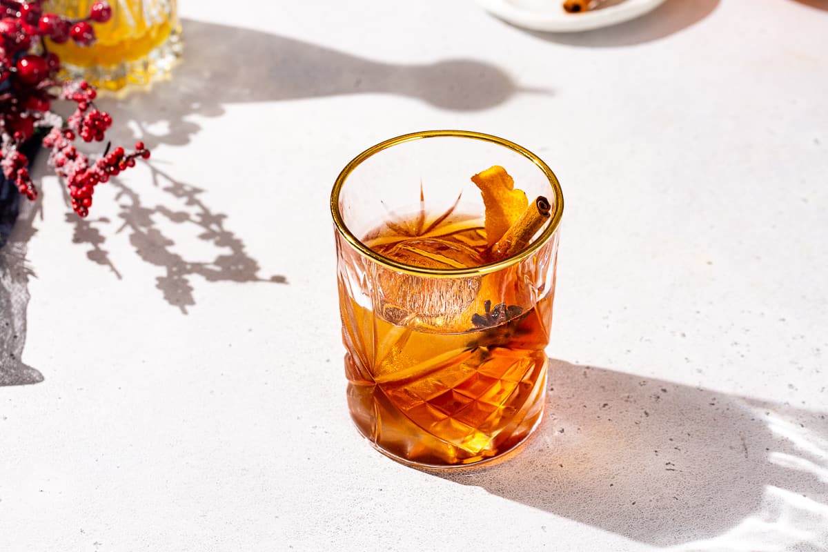 Slightly overhead view of the Christmas Old Fashioned cocktail on a countertop. The drink is in a gold-rimmed crystal glass with amber colored liquid and a large ice sphere. There is a star anise, cinnamon stick and orange peel garnish.