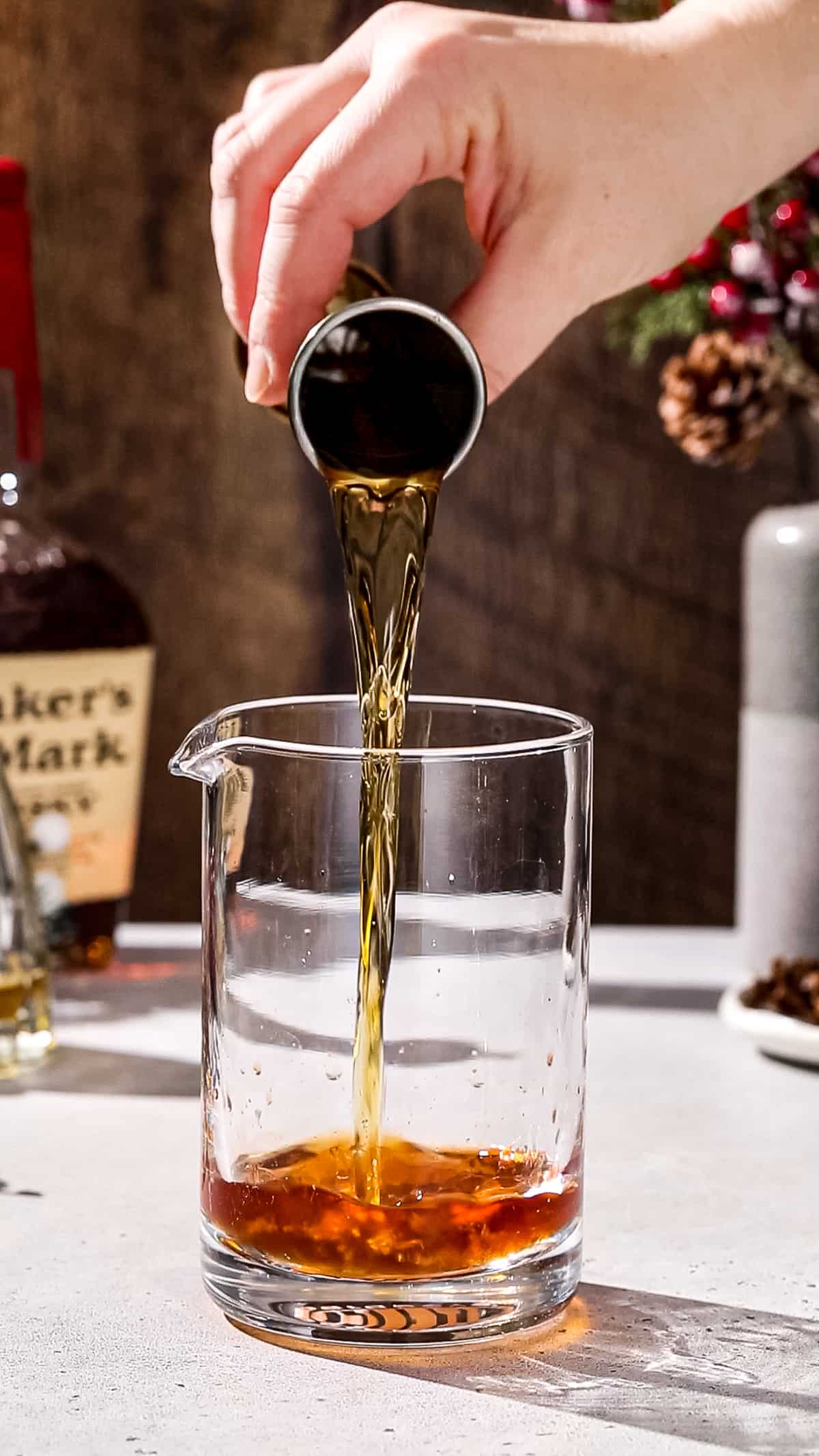 Hand using a gold jigger to add bourbon to a cocktail mixing glass.