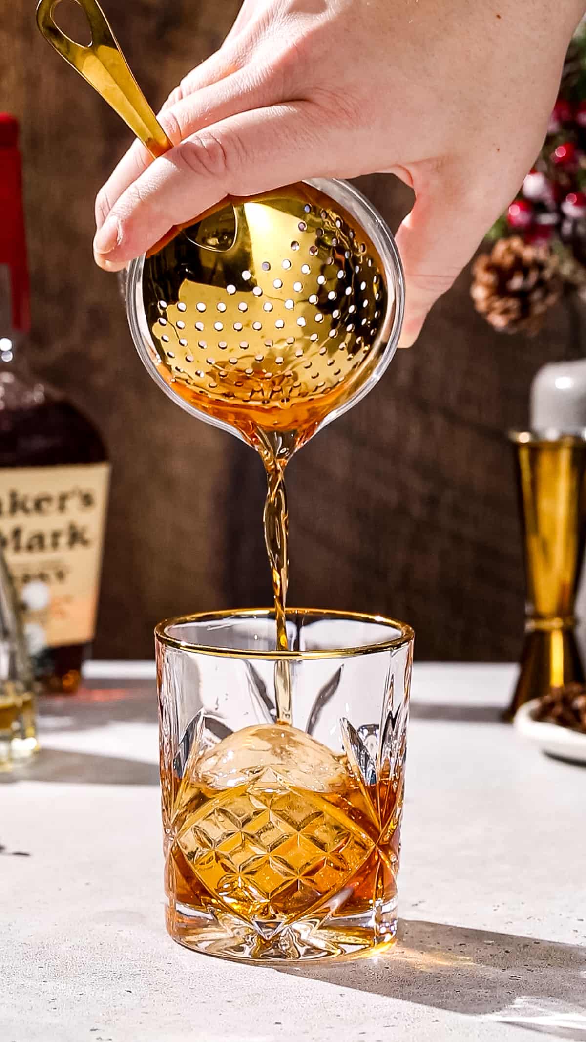 Hand using a gold julep strainer to strain a cocktail into a serving glass that has a large ice sphere in it.