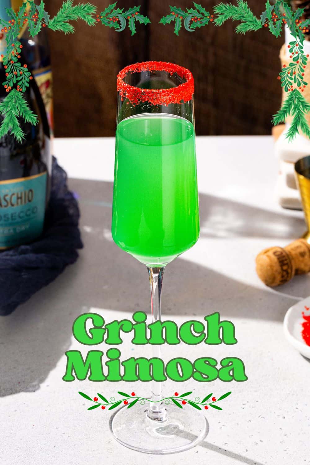 Grinch Mimosa on a countertop. The drink is bright green with red sugar around the rim. In the background is a bottle of Prosecco and a dish of red sugar. Bright green overlay text under the drink says “Grinch Mimosa”.