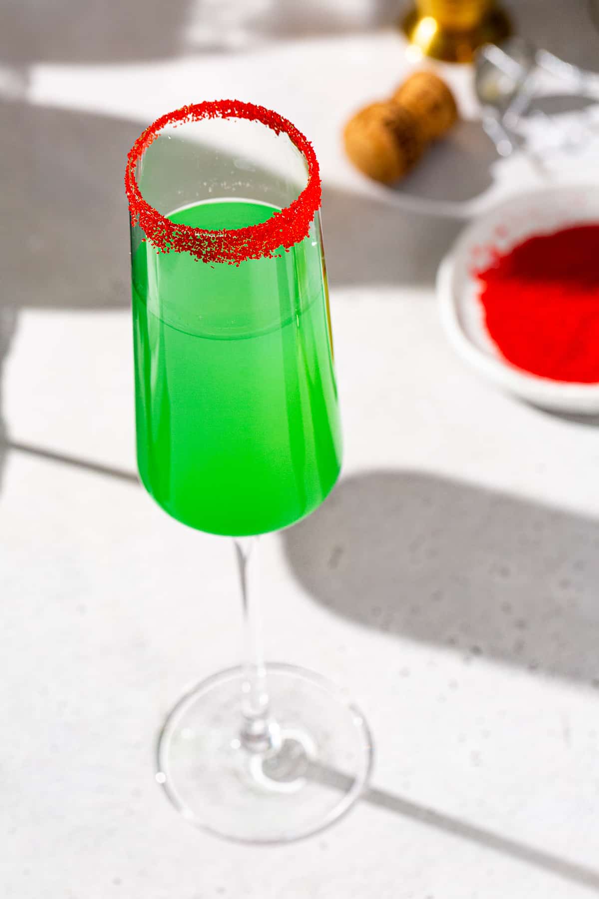 Overhead view of a Grinch Mimosa cocktail in a Champagne flute. A dish of red sugar and a champagne cork are in the background.