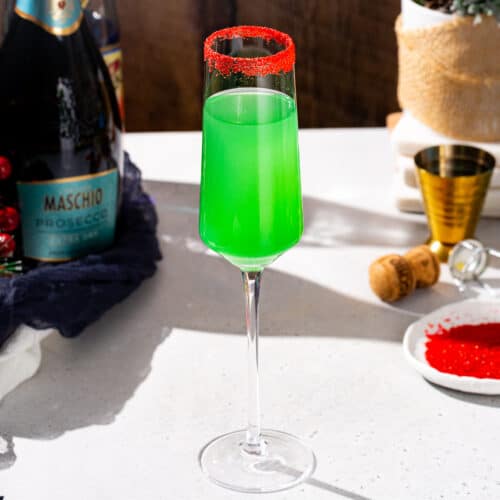 Side view of a green Grinch Mimosa cocktail with a red sugar rim in a Champagne flute. There is another cocktail in the background along with a jigger, a dish of red sugar and Prosecco.