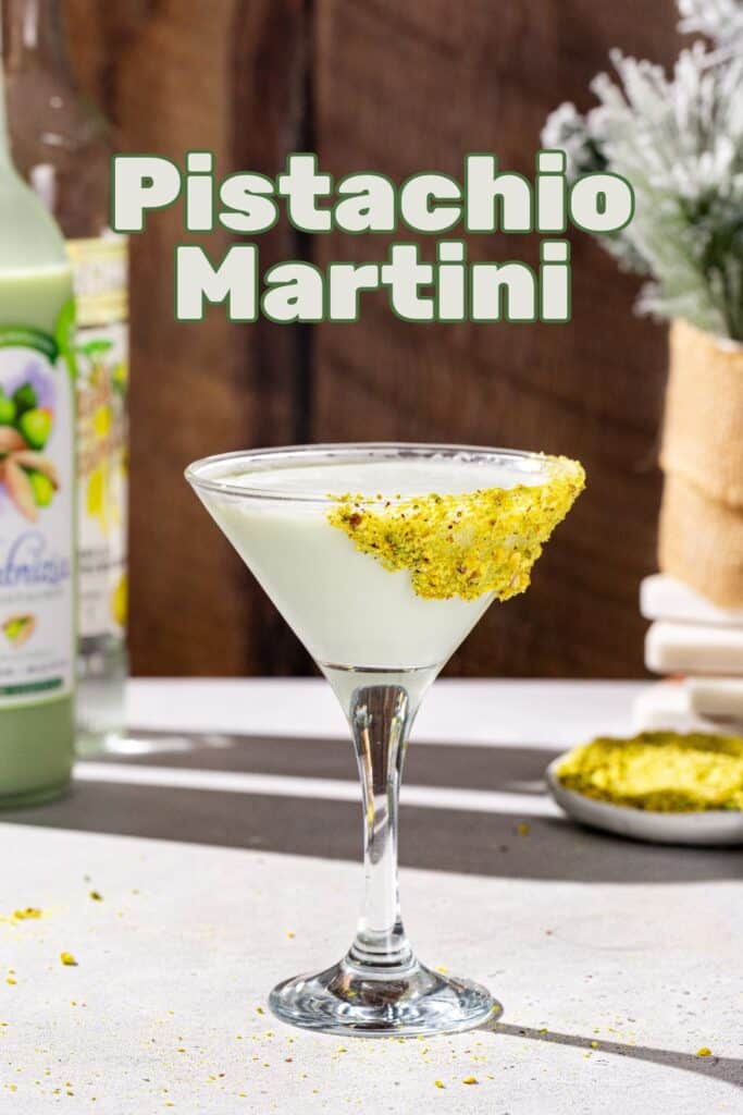 Side view of a Pistachio Martini cocktail in a martini glass. The drink is a pale green color and the glass is garnished with crushed up pistachios. In the background are a bottle of pistachio liqueur and vanilla vodka, plus some more crushed pistachios and an evergreen plant. Bold text at the top says “pistachio martini”.