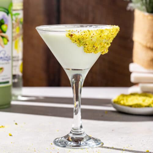 Side view of a Pistachio Martini cocktail in a martini glass. The drink is a pale green color and the glass is garnished with crushed up pistachios. In the background are a bottle of pistachio liqueur and vanilla vodka, plus some more crushed pistachios.