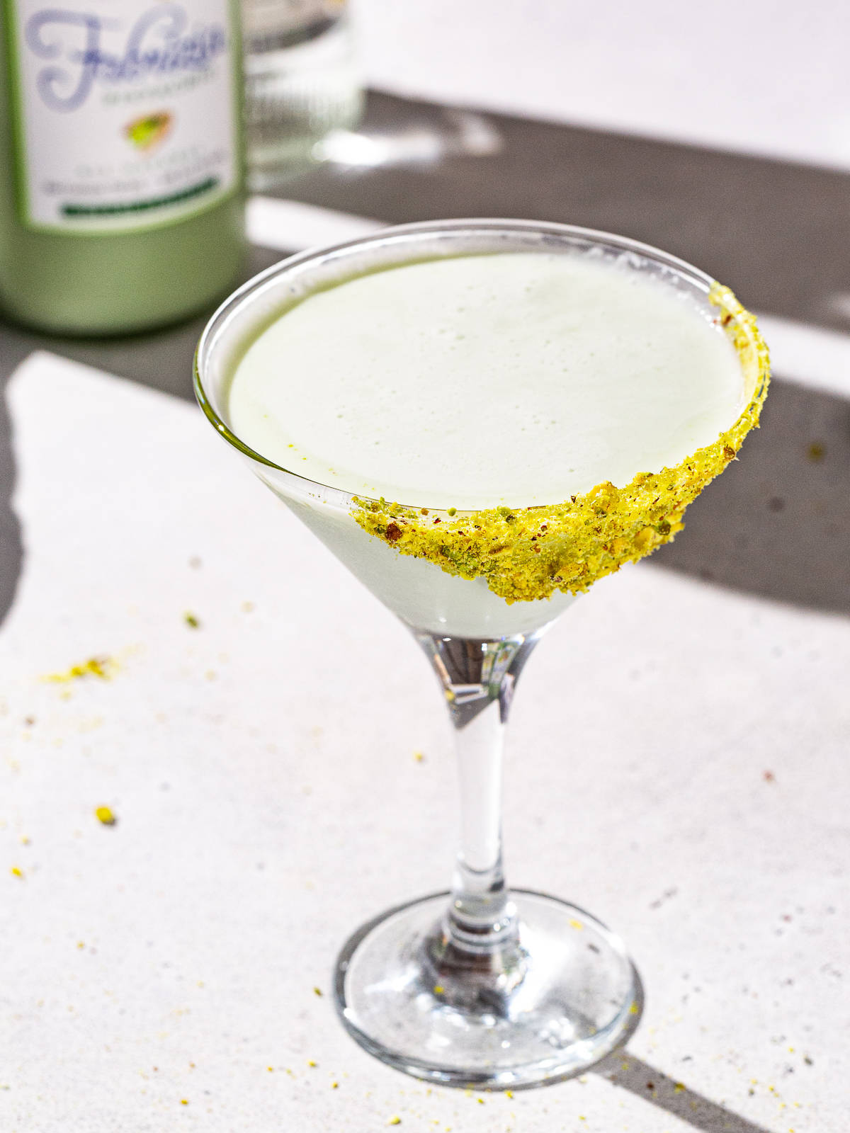 Overhead view of a pale green colored Pistachio Martini in a martini glass. In the background is a bottle of pistachio liqueur.