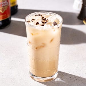 Slightly overhead view of a beige colored Smith and Kearns cocktail over ice in a tall glass. Chocolate curls are sprinkled on top of the drink. In the background are bottles of liqueur and a jigger.