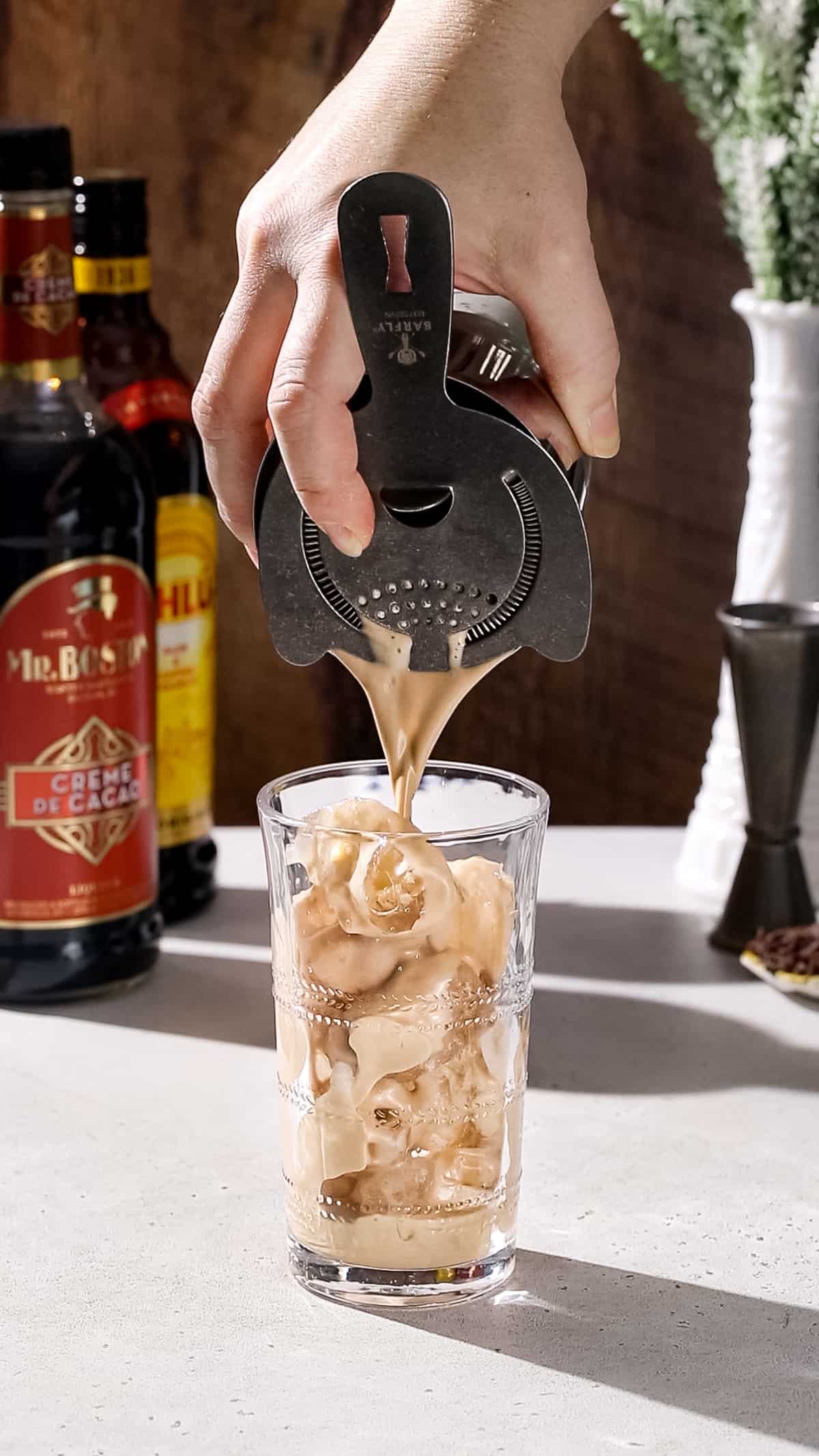 Hand straining a light brown liquid into a tall cocktail glass filled with ice.