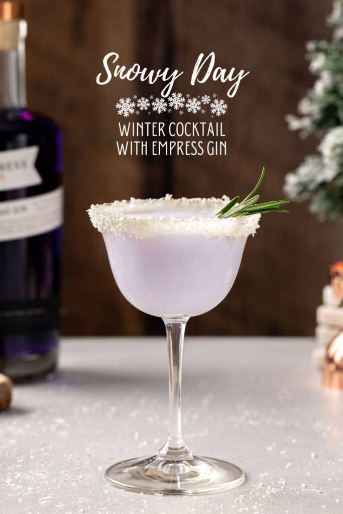 Snowy Day Cocktail in a coupe glass on a countertop. The drink is light purple in color and the rim of the glass is coated in dried coconut. A bottle of Empress gin and some Christmas decor is in the background. Above the drink, text overlay says “Snowy Day - winter cocktail with Empress Gin”.