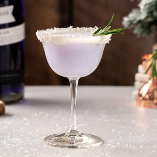 Side view of the Snowy Day cocktail, a winter gin cocktail with a light purple color. The rim is coated in dried coconut and a sprig of rosemary sticks out of the drink. In the background is a bottle of Empress gin, a jigger and a mini evergreen tree.