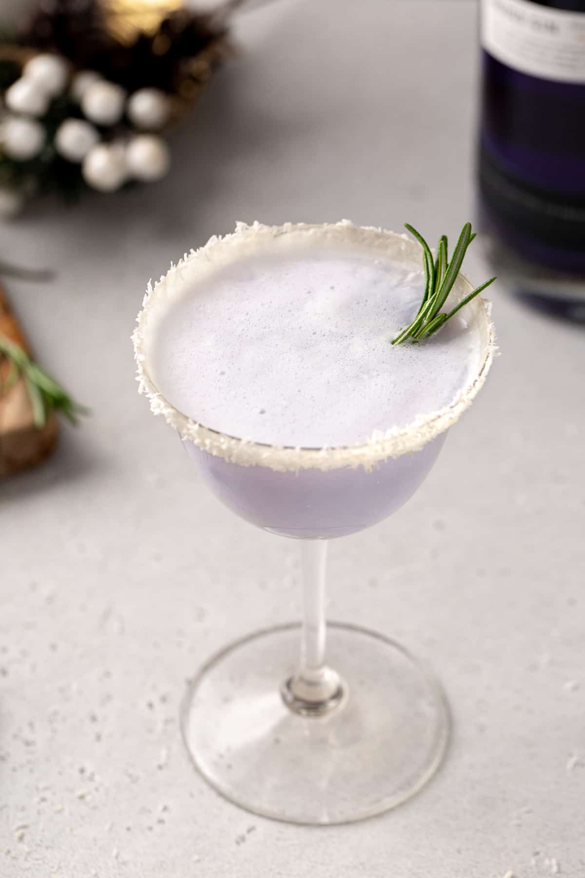 Overhead shot of the purple-colored Snowy Day cocktail, an Empress winter gin cocktail with coconut. A sprig of rosemary garnishes the drink.